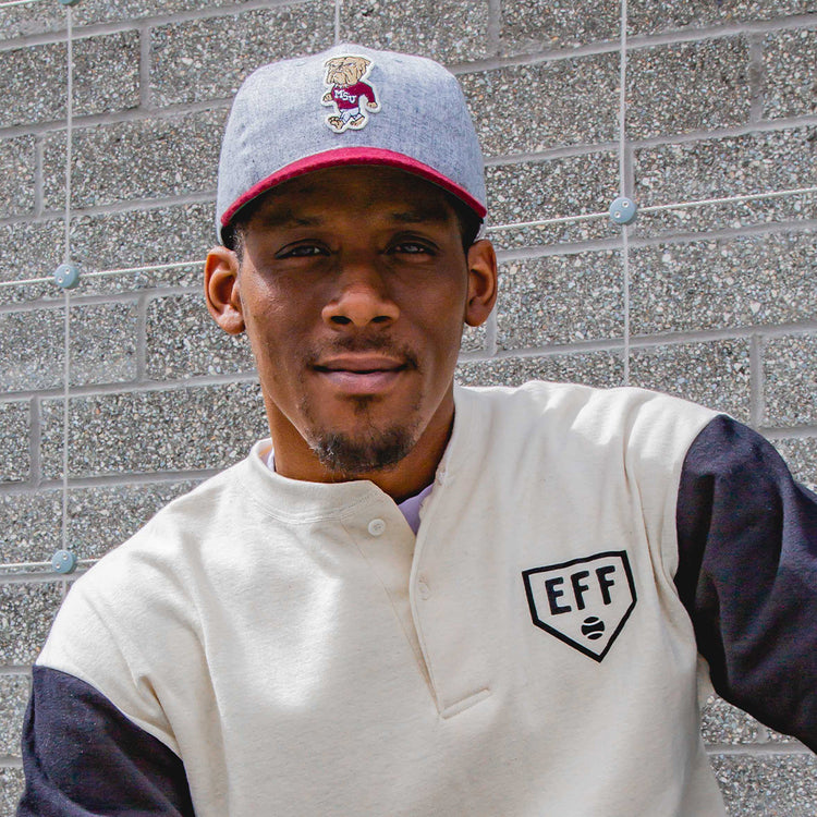 A man wearing the EFF Mississippi State University Mascot Vintage Ballcap and a white shirt with black sleeves sits in front of a gray brick wall, looking at the camera with a neutral expression.
