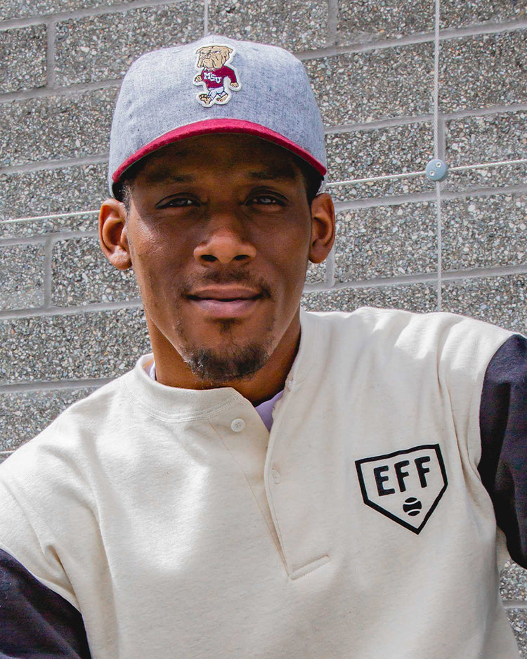 A man wearing the EFF Mississippi State University Mascot Vintage Ballcap and a white shirt with black sleeves sits in front of a gray brick wall, looking at the camera with a neutral expression.