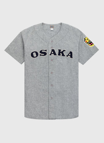 The Ebbets Field Flannels Osaka Tigers 1959 Authentic Wool Road Baseball Jersey features gray flannel, bold black OSAKA lettering, and a colorful tiger head patch on the sleeve, inspired by classic Koshien Field uniforms.