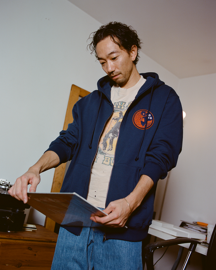Man wearing a navy hoodie with a Hiroshima Carp logo and jeans holding a record in a room.