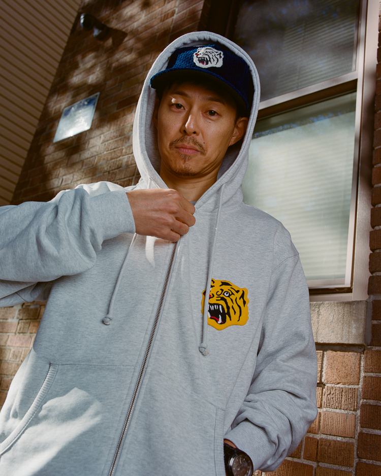 Person wearing a gray hoodie with an Osaka Tigers logo and an indigo corduroy cap with an Osaka Tigers logo, standing against a brick wall.