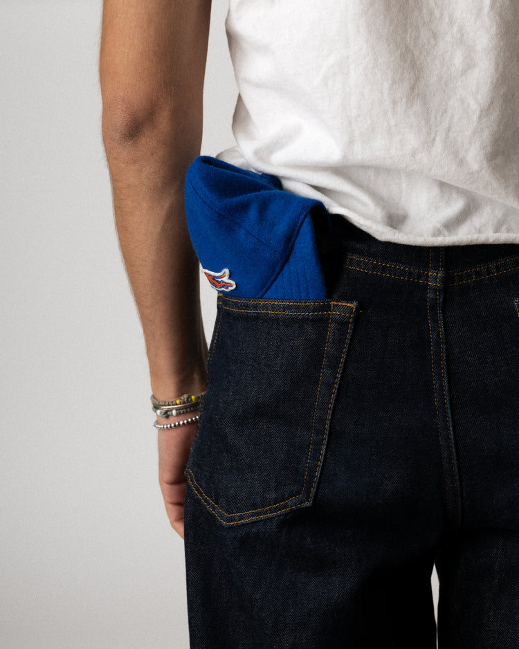 A person in a white t-shirt and dark jeans has an EFF Pacific Greyhounds 1939 Authentic Wool Softbrim Ballcap tucked into their back pocket, with several bracelets on their wrist.