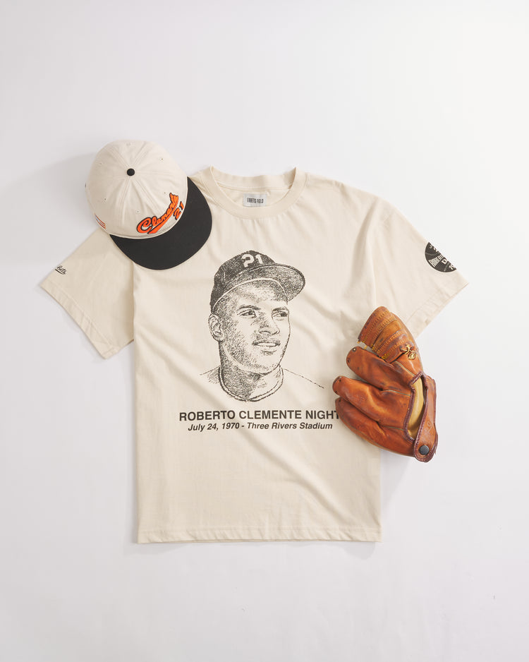 A cream-colored Ebbets Field “The Clemente Museum x Ebbets Field Short Sleeve Graphic T-Shirt,” featuring a Roberto Clemente portrait and event details, is arranged alongside a baseball glove and a white Orioles cap with a black brim on a white background, capturing the spirit of Roberto Clemente Day.