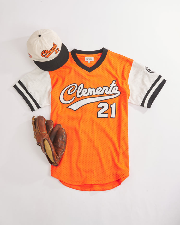 The Ebbets Field Clemente Museum x Ebbets Field V-Neck Mesh Jersey in bright orange, featuring “Clemente 21” on the front, is styled with a cream cap embroidered with “Clemente 21” and a brown leather baseball glove—all showcased on a white background for a truly standout Roberto Clemente jersey collection.