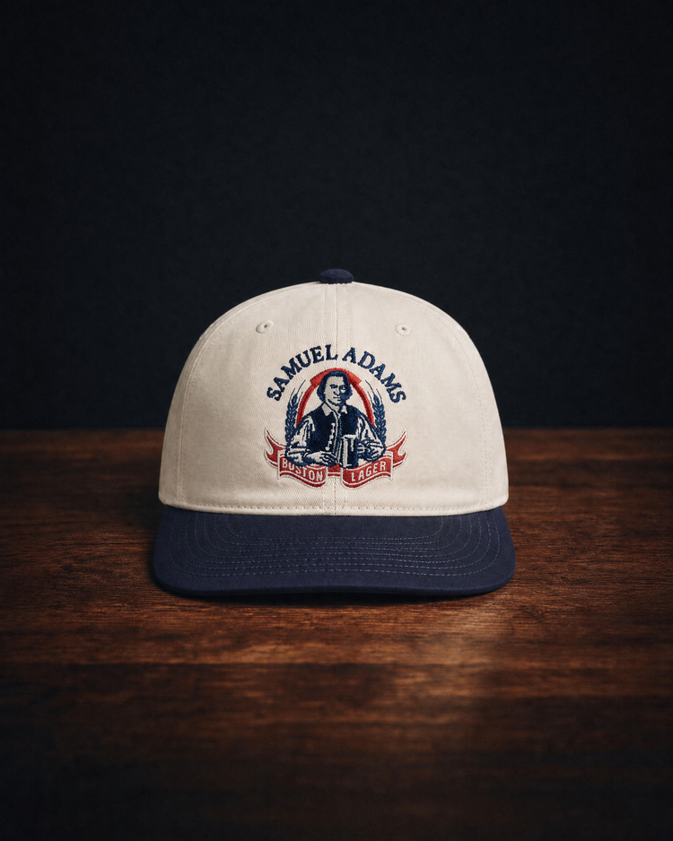 A beige and navy Sam Adams Crest Cotton Twill Ballcap by Ebbets Field, featuring the Samuel Adams Boston Lager vintage crest and a man holding a beer, rests on a wooden surface against a dark background.