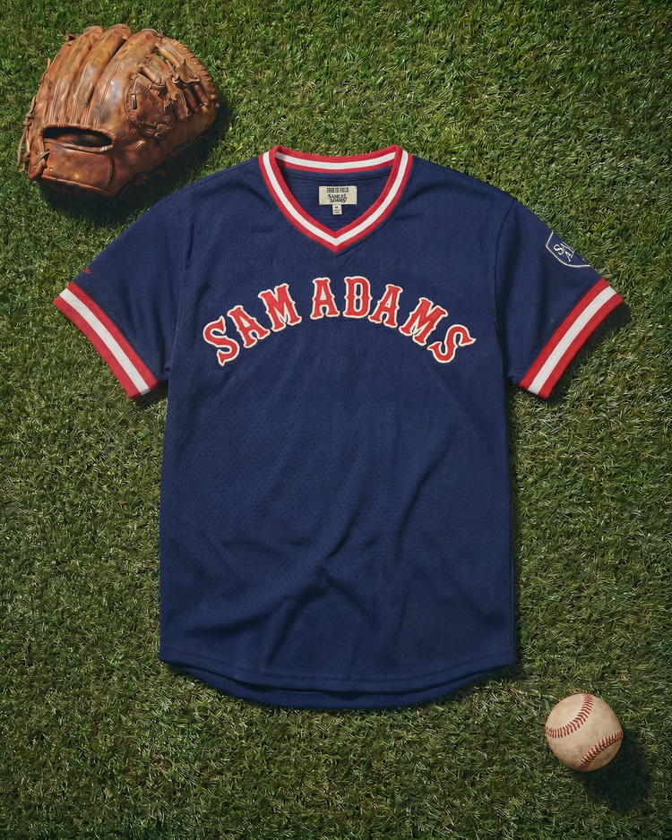 A Sam Adams Mesh V-Neck Jersey by Ebbets Field, in navy blue vintage style, is displayed on grass with a brown baseball glove and a white baseball with red stitching.