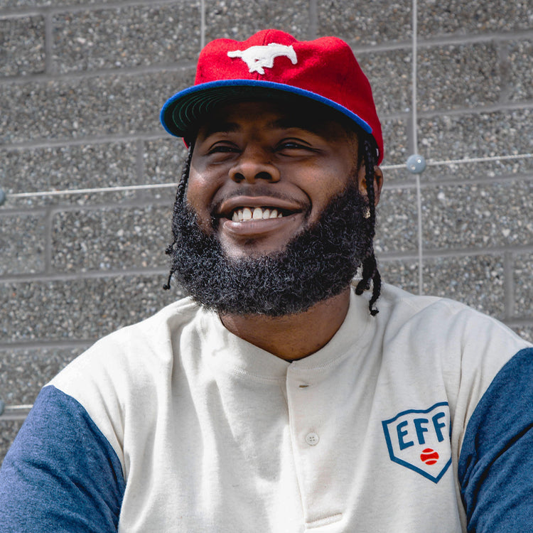 A man with a beard and braided hair smiles while wearing the EFF Southern Methodist University Mascot Vintage Ballcap and a cream shirt with blue sleeves, standing in front of a gray textured brick wall.