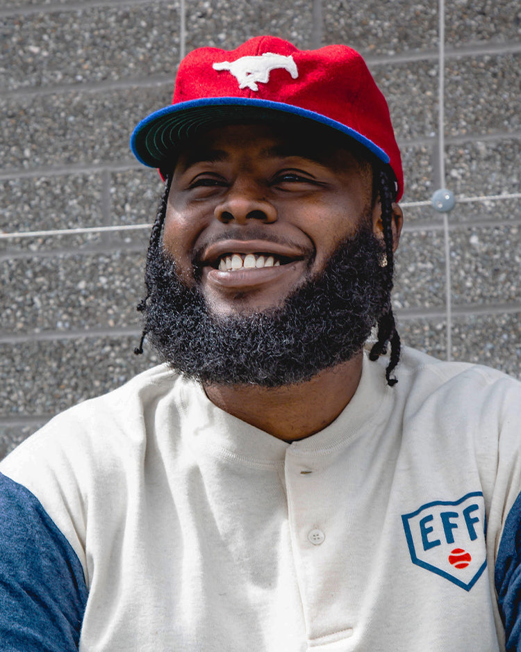 A man with a beard and braided hair smiles while wearing the EFF Southern Methodist University Mascot Vintage Ballcap and a cream shirt with blue sleeves, standing in front of a gray textured brick wall.