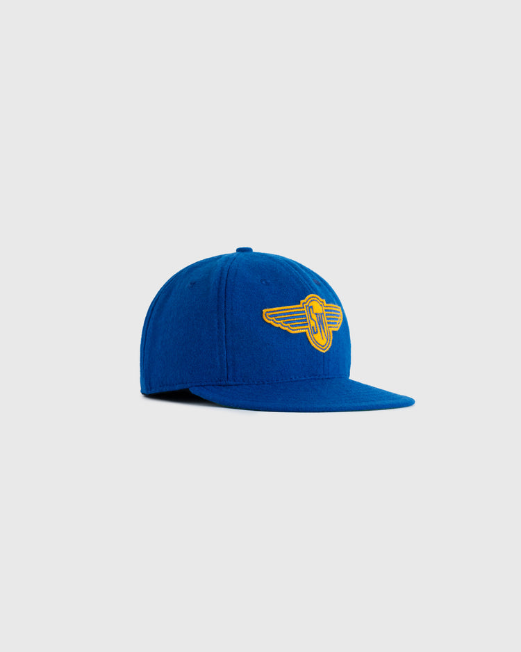 The EFF Stewart-Warner 1930 Authentic Wool Softbrim Ballcap features blue wool and a yellow embroidered winged logo, faithfully reproducing the original, shown against a plain light gray background.