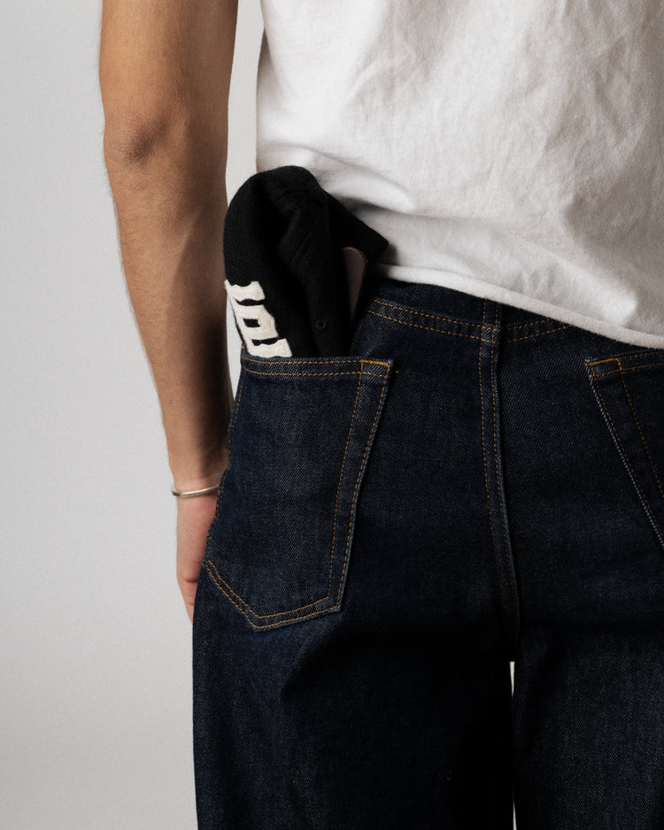 A person in dark blue jeans and a white t-shirt has a black EFF Tokyo Kyojin 1940 Authentic Wool Softbrim Ballcap with white lettering tucked into their back pocket; only the lower half of their back and one arm are visible.