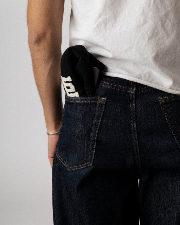 A person wearing a white t-shirt and dark blue jeans has a folded black item, possibly a hat or cloth with white lettering, sticking out of their back pocket. Only the lower back and one arm are visible.