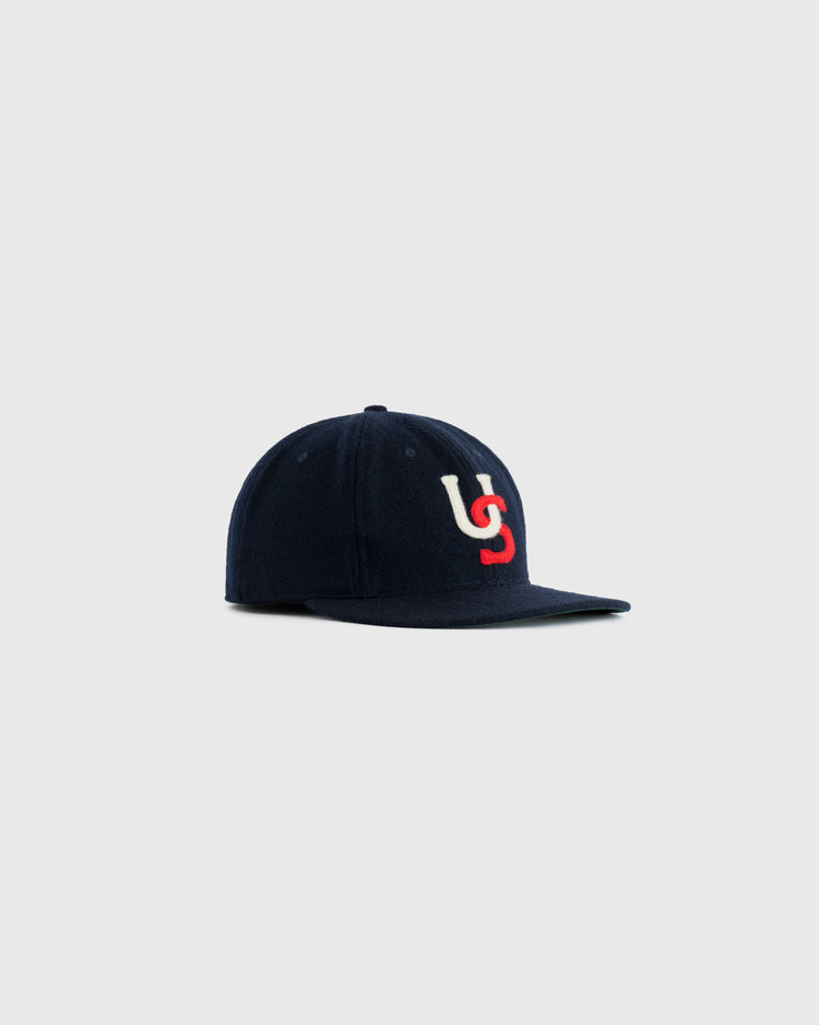 The Ebbets Field Flannels US All-Stars 1957 Authentic Wool Ballcap is a navy blue cap with a white and red US logo, inspired by the 1957 All-Stars ballcap, shown on a plain light gray background.