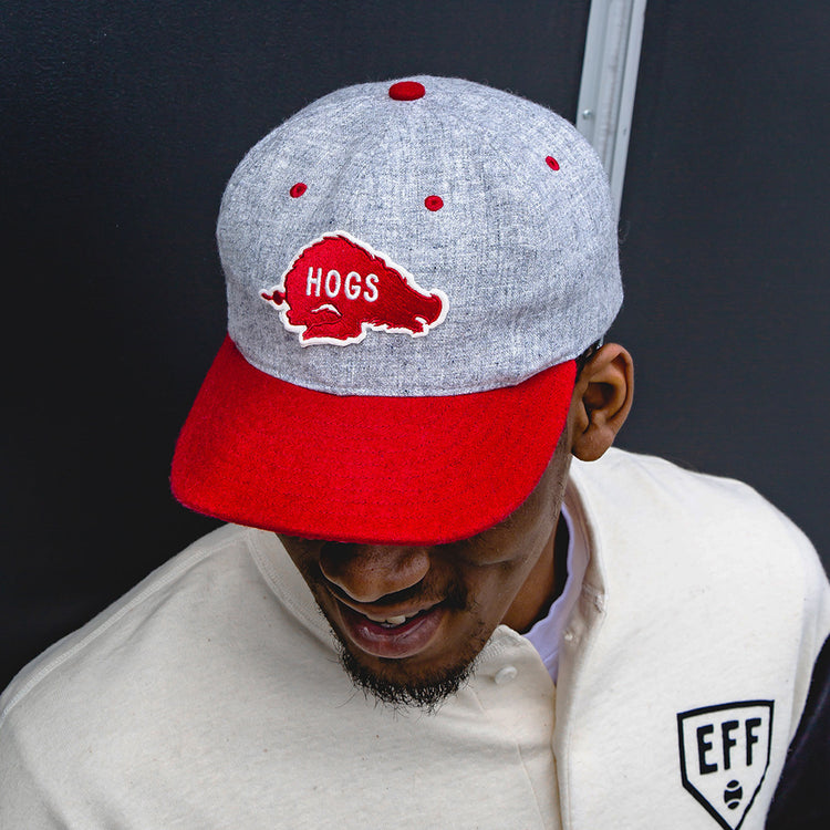 A man wearing a University of Arkansas Mascot Vintage Ballcap by EFF, featuring a red hog and HOGS on the front, and a cream jacket with a black EFF patch, smiles and looks downward against a dark background.