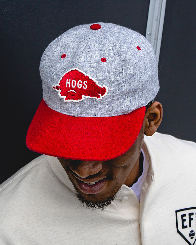 A man wearing a University of Arkansas Mascot Vintage Ballcap by EFF, featuring a red hog and HOGS on the front, and a cream jacket with a black EFF patch, smiles and looks downward against a dark background.