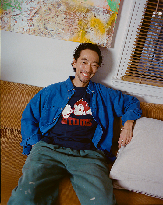 A smiling man with wavy hair sits on a tan couch, wearing a blue shirt over an Ebbets Field Sankei Atoms Short Sleeve Graphic Tee. In the background are a painting and window blinds, adding a subtle nod to Japanese baseball style.