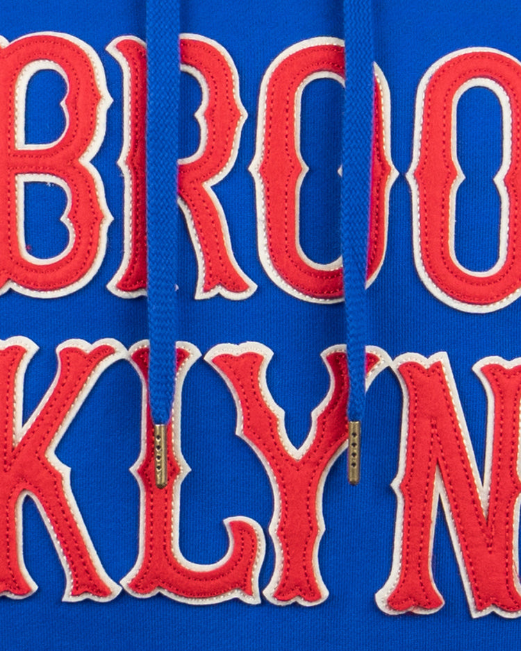 Close-up of EFF Brooklyn Royal Giants French Terry Script Hooded Sweatshirt: blue fabric with red and white BROO KLYN text, plus a matching blue drawstring with metal tips down the center.