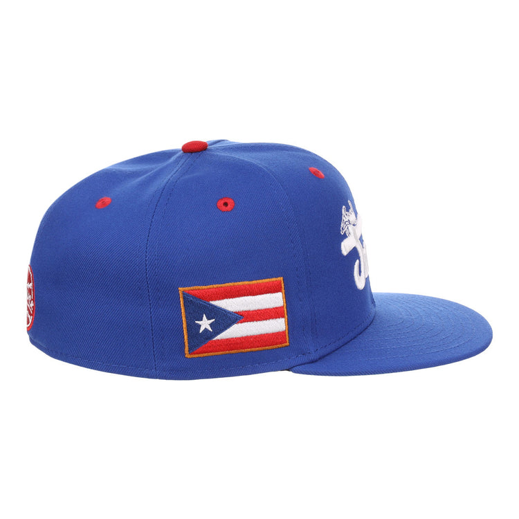 The EFF Santurce Cangrejeros NLB Flip Fitted Ballcap is a blue cap with a white embroidered front design, Puerto Rican flag patch on the side, red top button detail, and an emblem on the back—honoring legends like Roberto Clemente.