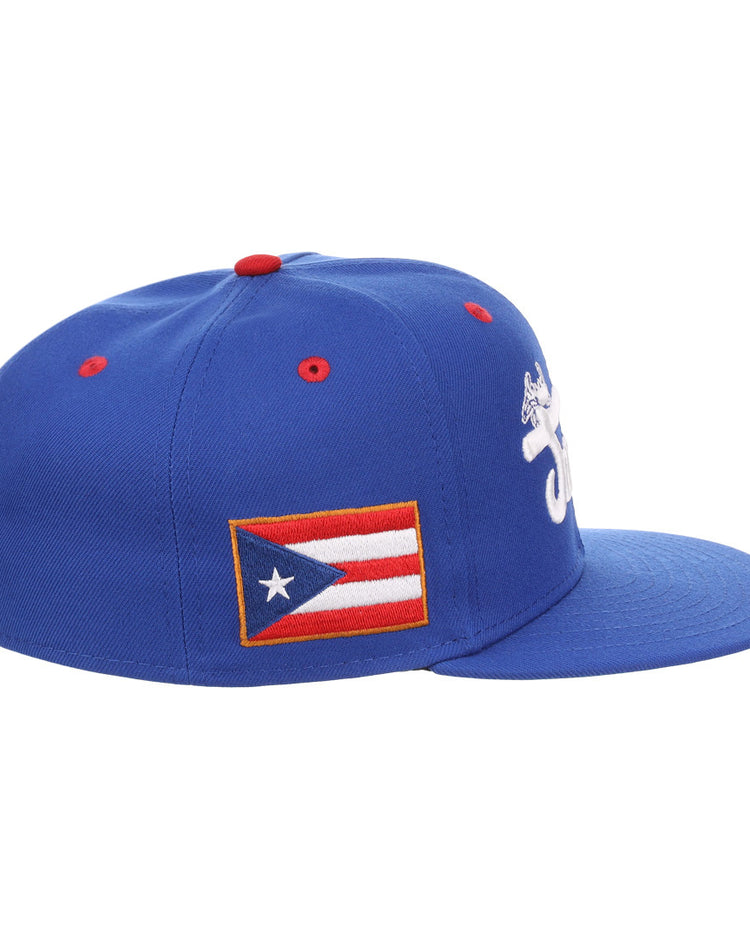 The EFF Santurce Cangrejeros NLB Flip Fitted Ballcap is a blue cap with a white embroidered front design, Puerto Rican flag patch on the side, red top button detail, and an emblem on the back—honoring legends like Roberto Clemente.