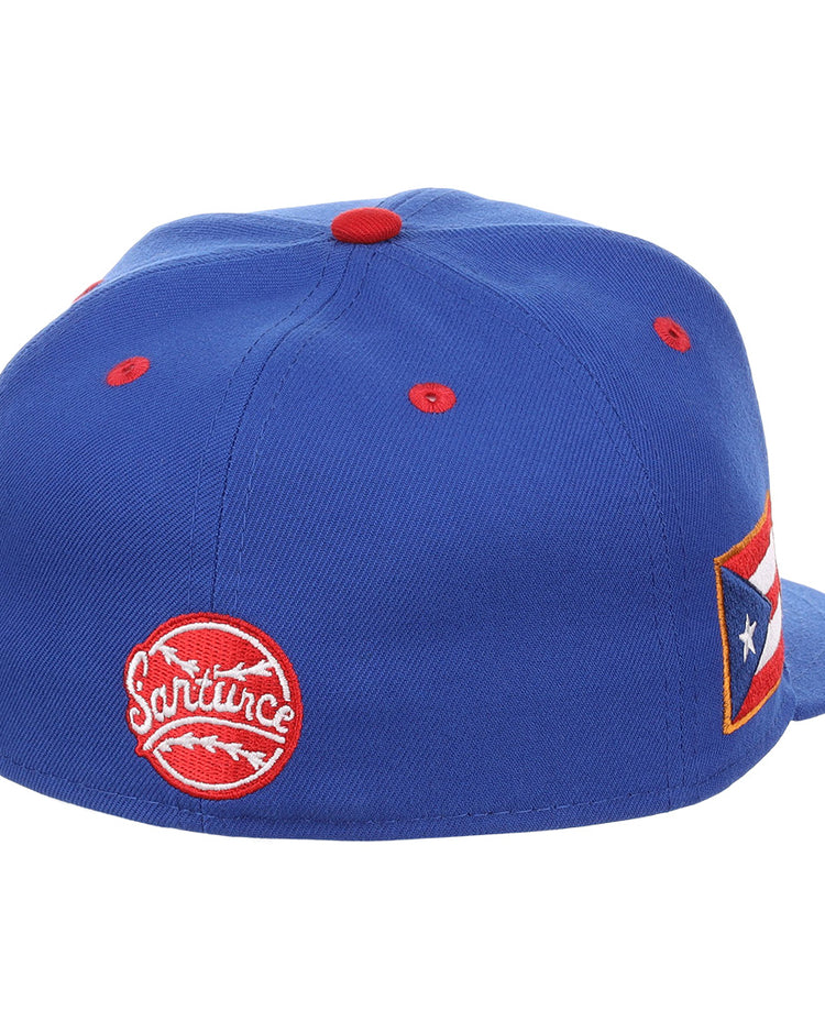 The EFF Santurce Cangrejeros NLB Flip Fitted Ballcap is a blue cap with red accents, a circular Santurce patch on the back, a Puerto Rican flag patch on the side, and is inspired by Puerto Rican League legends like Roberto Clemente.