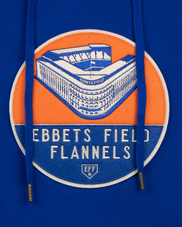 The EFF Ebbets Field Flannels French Terry Script Hooded Sweatshirt is blue, featuring a circular Ebbets Field patch on an orange background with blue drawstrings partially covering the design—a nod to authentic historical sportswear.