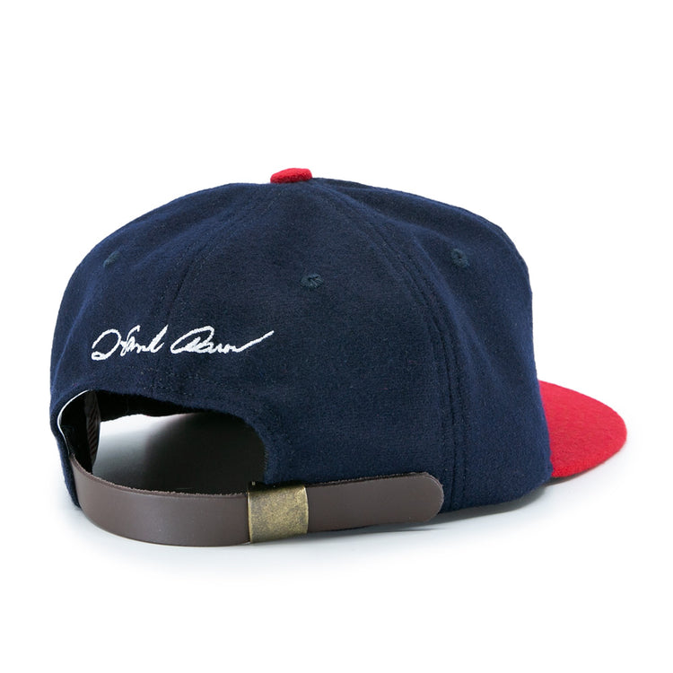The Ebbets Field Flannels Hank Aaron 1957 Signature Series Ballcap features navy wool, a red brim, a brown leather strap with brass clasp, white cursive embroidery near the back opening, and is proudly made in the USA.