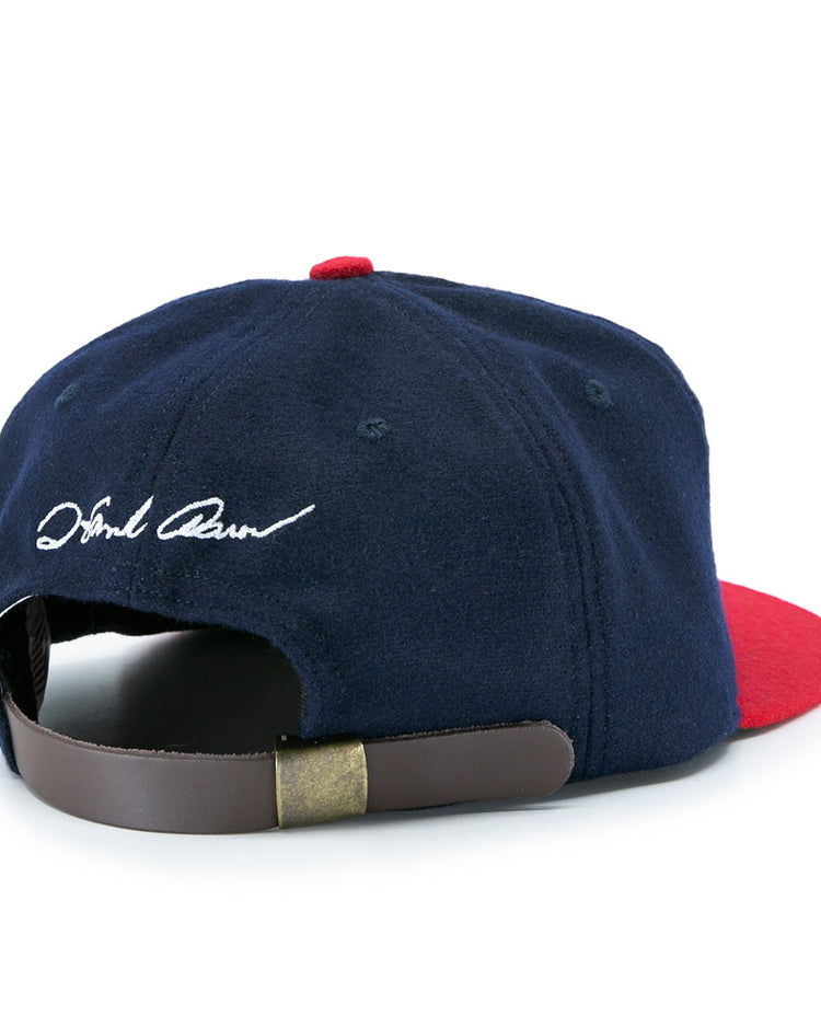 The Ebbets Field Flannels Hank Aaron 1957 Signature Series Ballcap features navy wool, a red brim, a brown leather strap with brass clasp, white cursive embroidery near the back opening, and is proudly made in the USA.