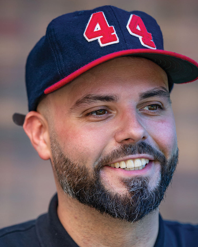 A bearded man smiles while wearing the Hank Aaron 1957 Signature Series Ballcap by Ebbets Field Flannels—a navy wool cap with red trim, the number 44, and Hank Aaron’s signature. The background is blurred.