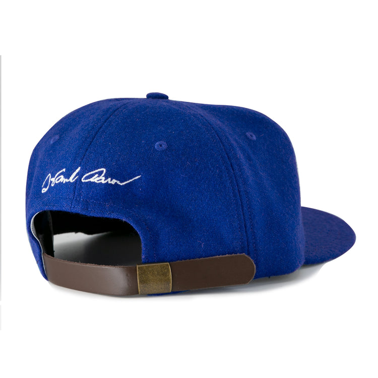 The Ebbets Field Flannels Hank Aaron 1973 Signature Series Ballcap features a royal blue wool design, white embroidered signature above an adjustable brown leather strap with brass buckle, and is proudly made in the USA.