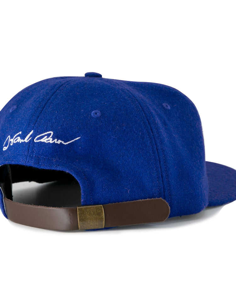 The Ebbets Field Flannels Hank Aaron 1973 Signature Series Ballcap features a royal blue wool design, white embroidered signature above an adjustable brown leather strap with brass buckle, and is proudly made in the USA.