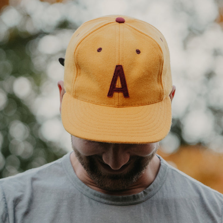 A man in a light gray shirt stands outdoors with blurred greenery behind him, looking down and partially obscuring his face while wearing the EFF Arizona State University 1955 Vintage Ballcap featuring a large maroon letter A.