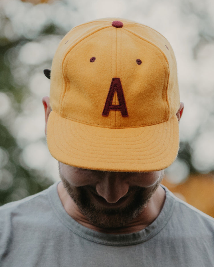 A man in a light gray shirt stands outdoors with blurred greenery behind him, looking down and partially obscuring his face while wearing the EFF Arizona State University 1955 Vintage Ballcap featuring a large maroon letter A.
