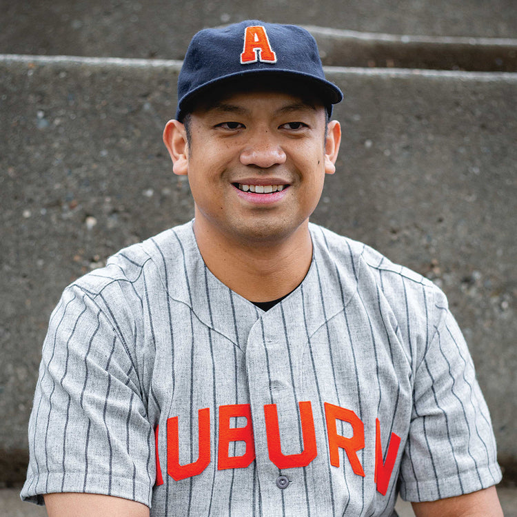 A smiling man wears the Auburn University 1966 Vintage Ballcap by Ebbets Field Flannels, featuring an orange A, along with a gray pinstriped jersey reading AUBURN in red, while seated in front of concrete steps.