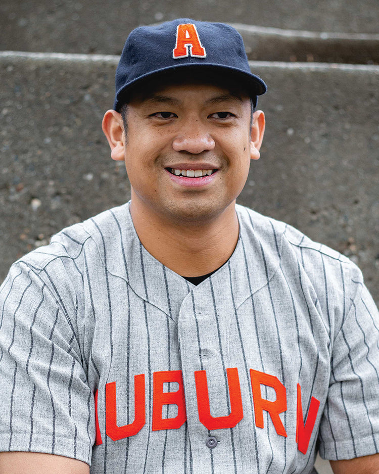 A smiling man wears the Auburn University 1966 Vintage Ballcap by Ebbets Field Flannels, featuring an orange A, along with a gray pinstriped jersey reading AUBURN in red, while seated in front of concrete steps.