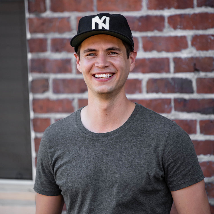 A smiling man wears the EFF Brooklyn Eagles Vintage Inspired Ballcap and a gray t-shirt, standing in front of a red brick wall.