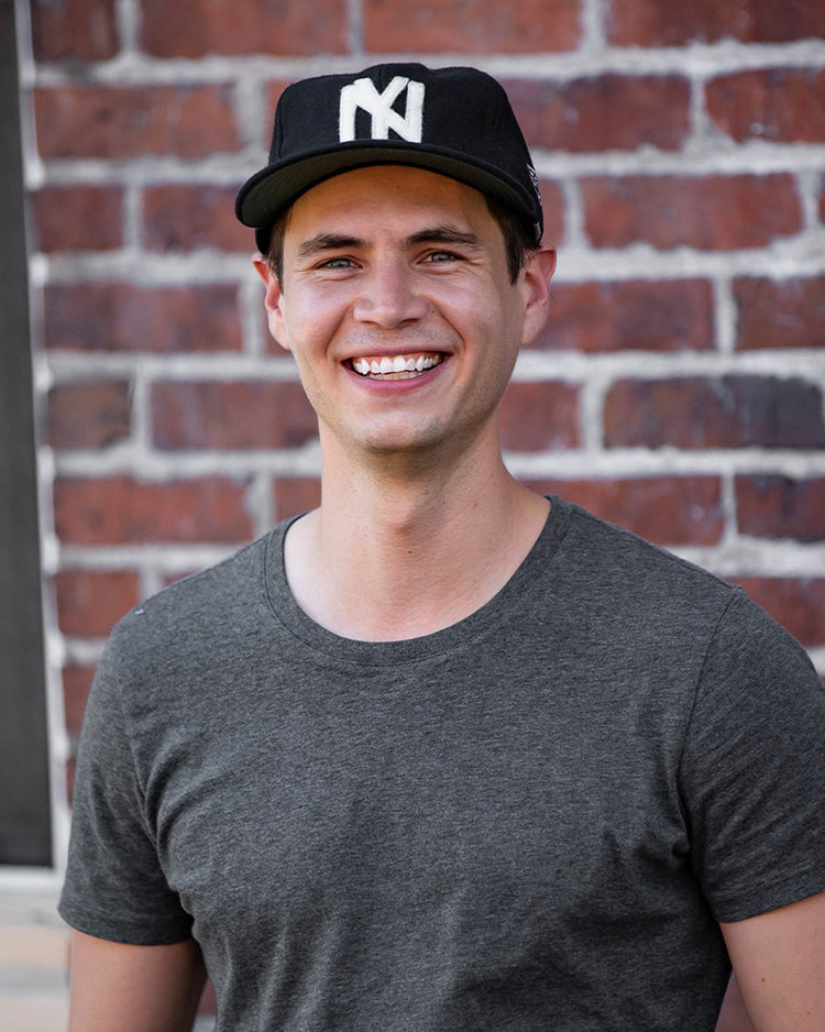 A smiling man wears the EFF Brooklyn Eagles Vintage Inspired Ballcap and a gray t-shirt, standing in front of a red brick wall.