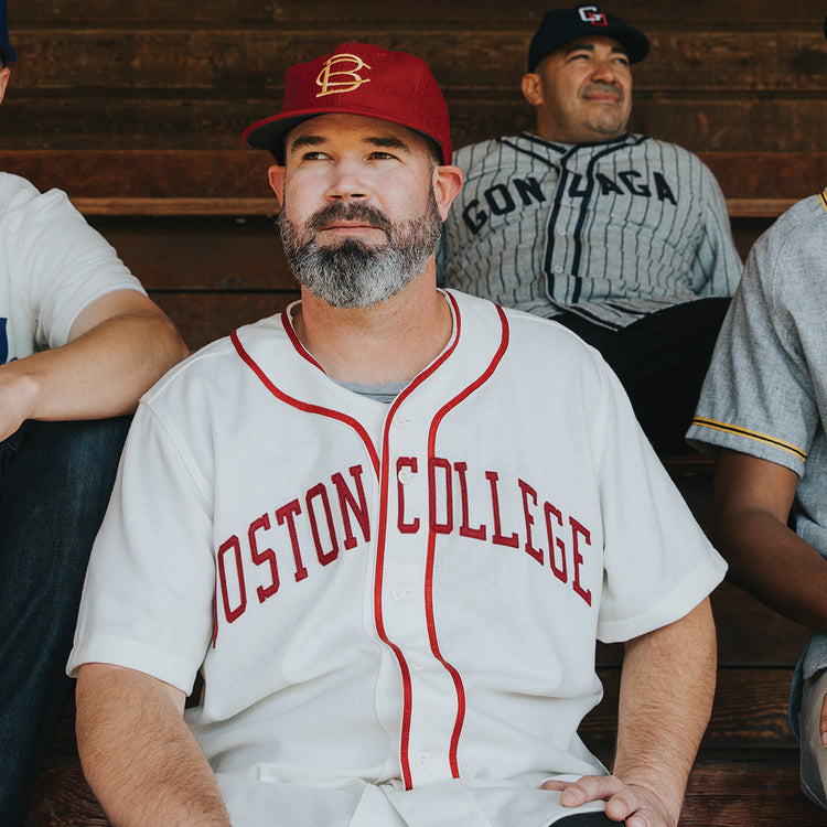 A man with a gray beard wears an EFF Boston College 1966 Vintage Ballcap in red and a white Boston College baseball jersey as he sits on wooden bleachers, surrounded by others in baseball jerseys.