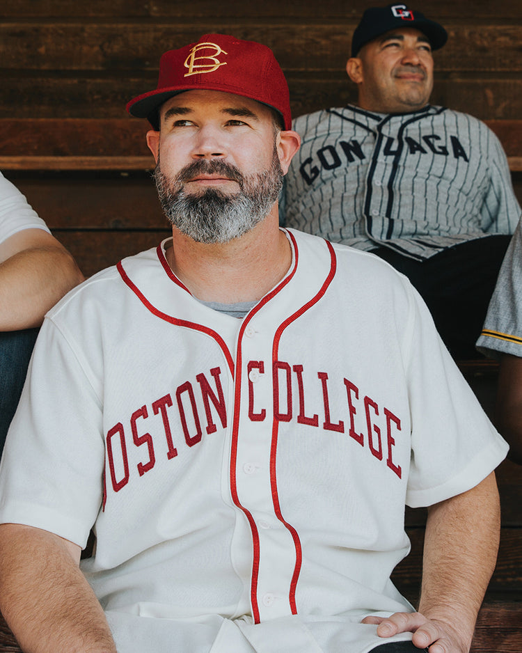 A man with a gray beard wears an EFF Boston College 1966 Vintage Ballcap in red and a white Boston College baseball jersey as he sits on wooden bleachers, surrounded by others in baseball jerseys.
