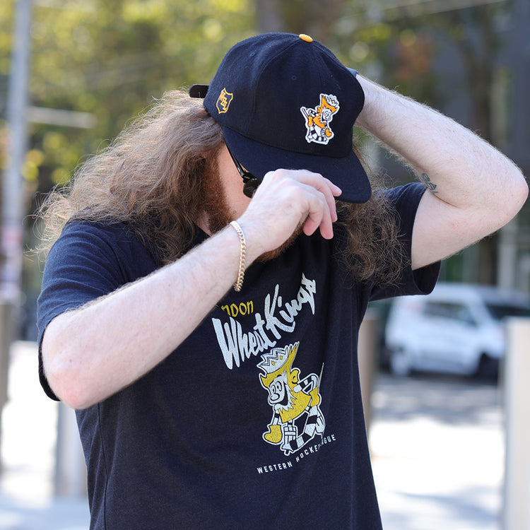 A person with long hair and a beard adjusts the EFF Brandon Wheat Kings 1955 Vintage Ballcap, featuring cartoon designs, partially covering their face. They pair it with a matching black T-shirt and gold bracelet while outdoors on a sunny day.