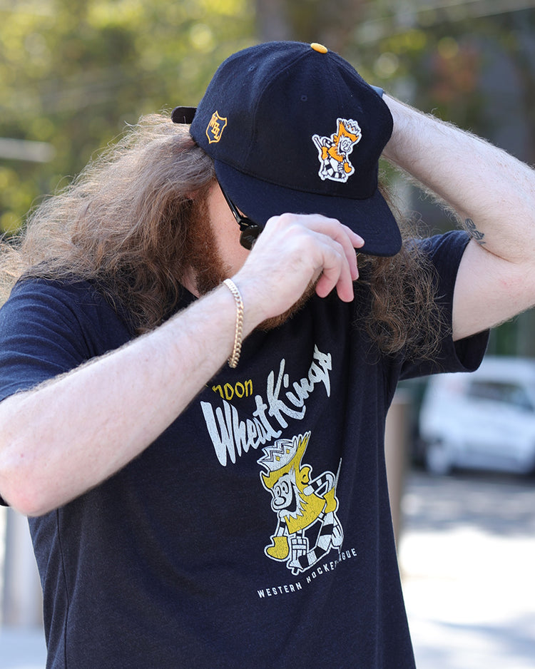 A person with long hair and a beard adjusts the EFF Brandon Wheat Kings 1955 Vintage Ballcap, featuring cartoon designs, partially covering their face. They pair it with a matching black T-shirt and gold bracelet while outdoors on a sunny day.
