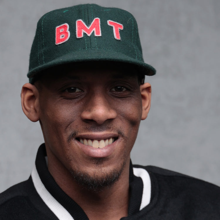 A smiling man wears the EFF Brooklyn Manhattan Transit Company 1939 Vintage Ballcap—a dark green wool baseball cap with red letters—and a black jacket with white trim, posing before a plain gray background.