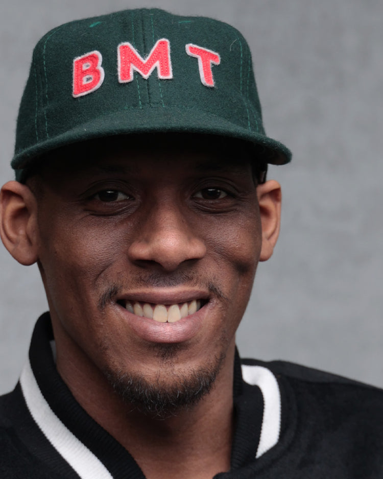 A smiling man wears the EFF Brooklyn Manhattan Transit Company 1939 Vintage Ballcap—a dark green wool baseball cap with red letters—and a black jacket with white trim, posing before a plain gray background.