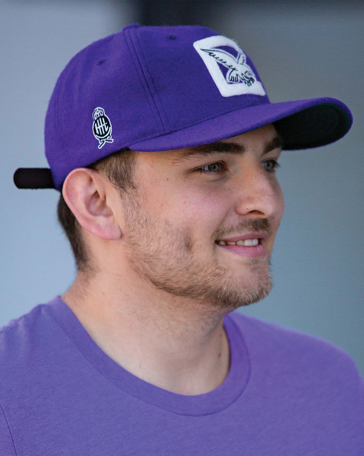 A young man with short brown hair and light stubble smiles while wearing the EFF Cleveland Falcons 1936 Vintage Ballcap—a purple wool cap with a bird logo and adjustable leather strap—standing in front of a blurred background.