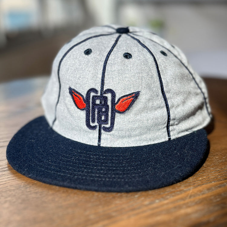 The EFF Columbia Park 1909 Vintage 8-Panel Ballcap in gray and navy, with a dark brim and embroidered logo of red wings and blue lines, sits on a wooden surface.