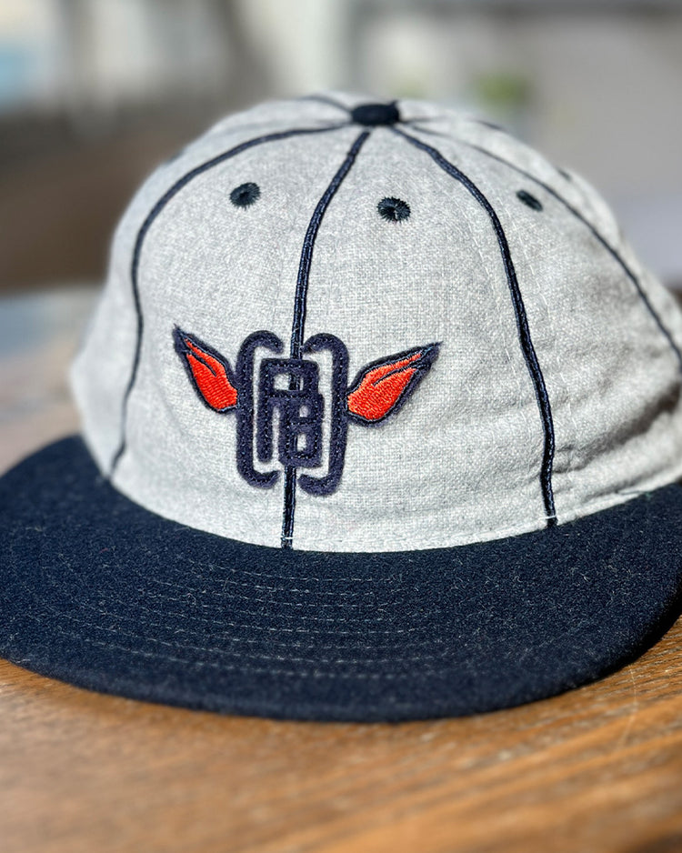 The EFF Columbia Park 1909 Vintage 8-Panel Ballcap in gray and navy, with a dark brim and embroidered logo of red wings and blue lines, sits on a wooden surface.