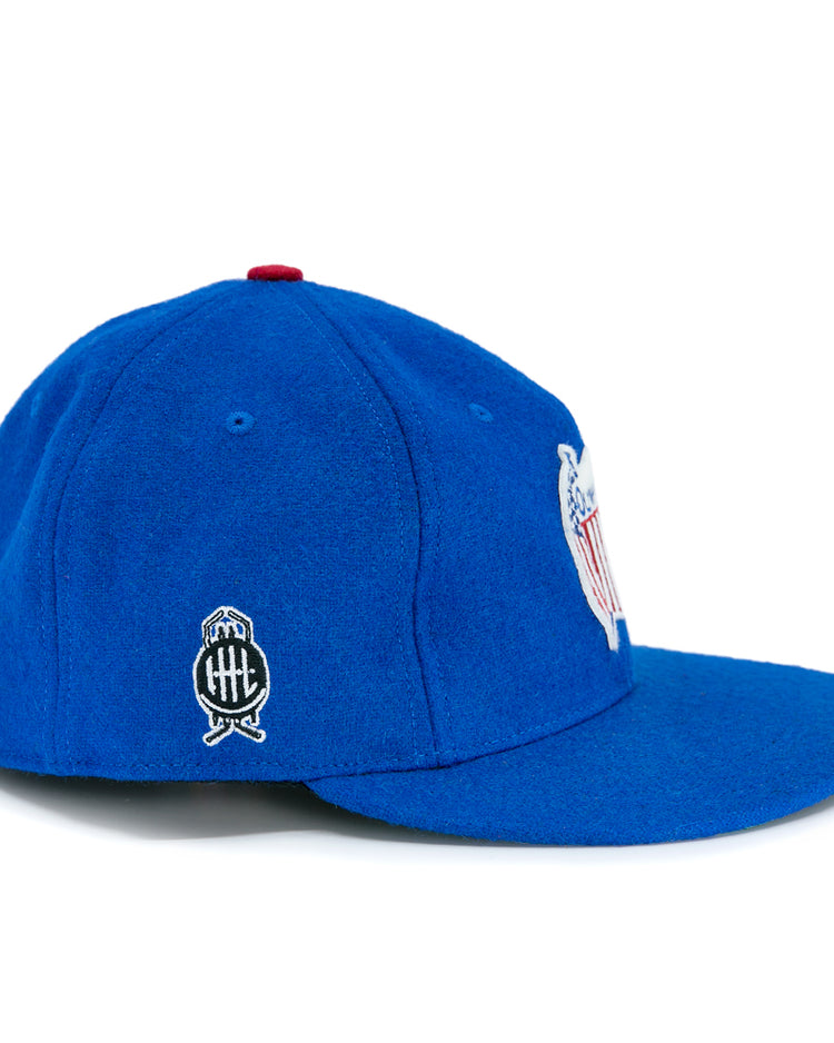 The Detroit Olympics Vintage Ballcap by EFF is a royal blue wool cap with a slightly curved brim, white and black side logo, red top button, and an adjustable leather strap for the perfect fit.