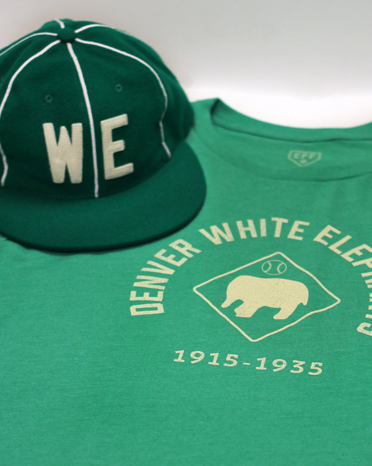 A green EFF Denver White Elephants T-Shirt (1915-1935 tribute) with a white elephant logo and diamond background sits beside a green cap featuring white WE letters and piping, honoring Negro Leagues baseball.