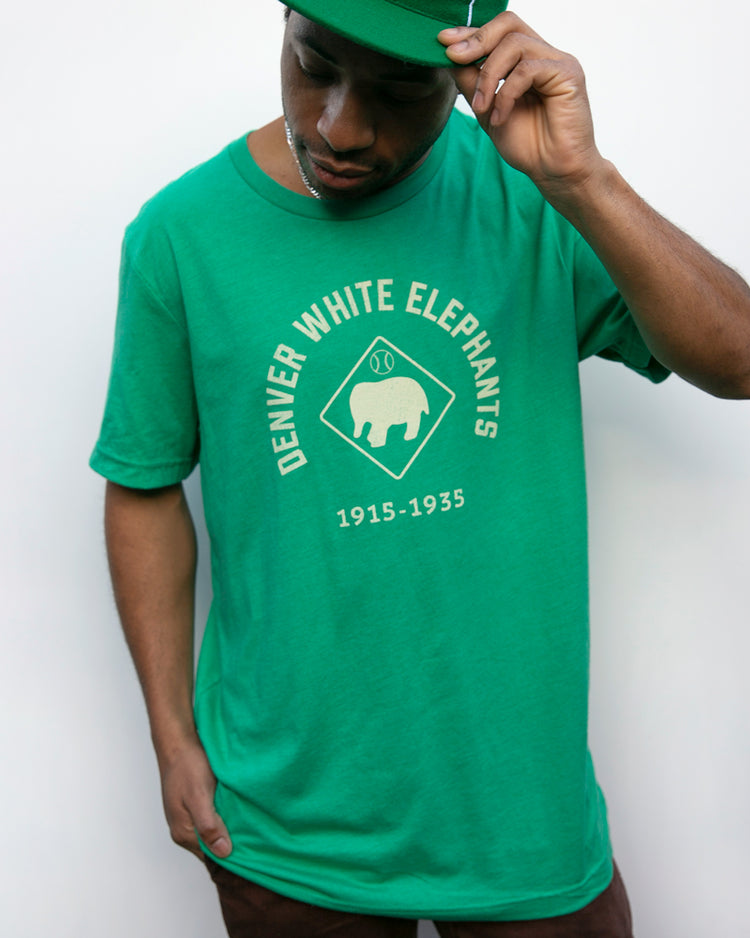 A man in a green Denver White Elephants T-Shirt by EFF adjusts his cap. The shirt features a white elephant graphic inside a diamond shape, honoring the Negro Leagues baseball team from 1915-1935.