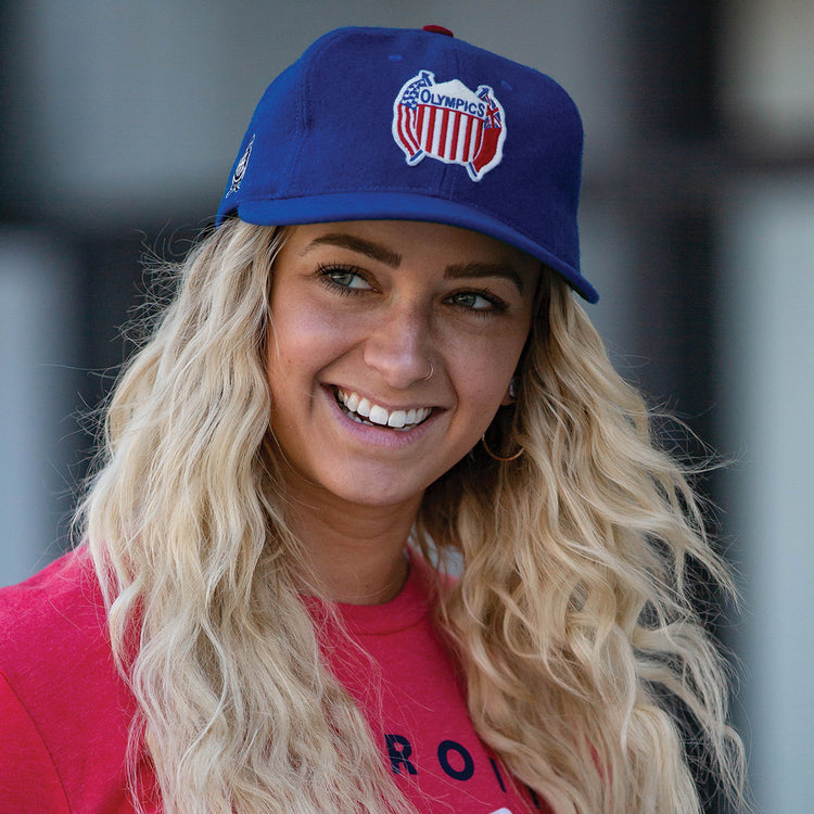 A smiling person with long, wavy blonde hair wears the EFF Detroit Olympics Vintage Ballcap, featuring a red, white, and blue Olympics logo. They pair it with a red shirt, set against a blurred background.