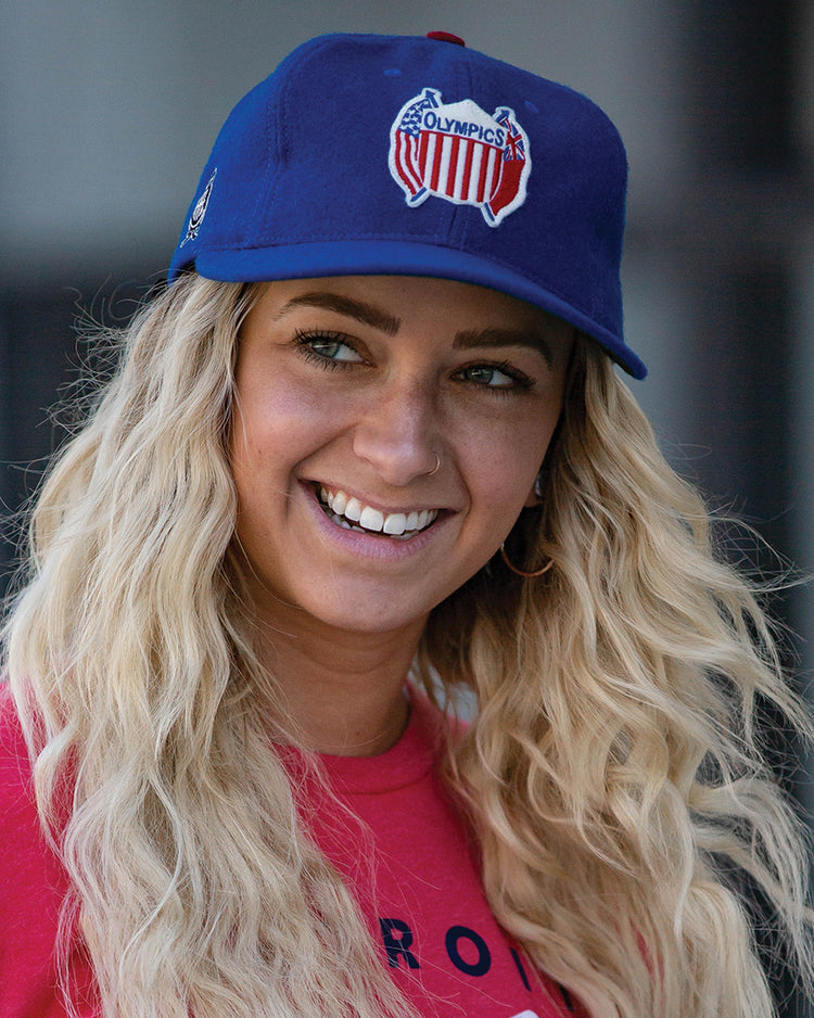 A smiling person with long, wavy blonde hair wears the EFF Detroit Olympics Vintage Ballcap, featuring a red, white, and blue Olympics logo. They pair it with a red shirt, set against a blurred background.