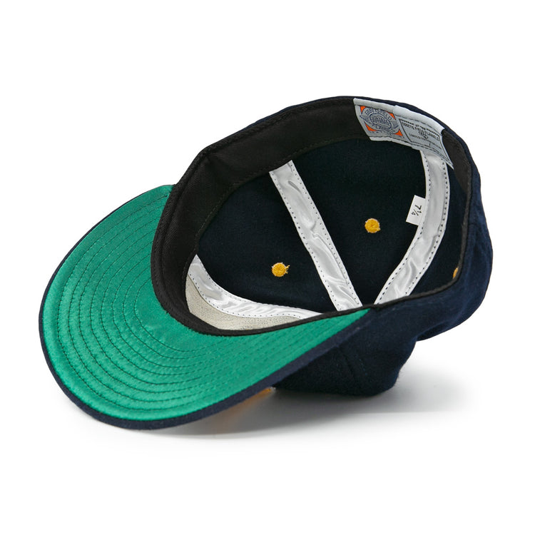 An authentic EFF Great Lakes Naval Station 1944 Vintage Ballcap, navy wool, is shown upside down to reveal its green underside brim, white lining, yellow rivets, visible labels, and size tag inside the crown.